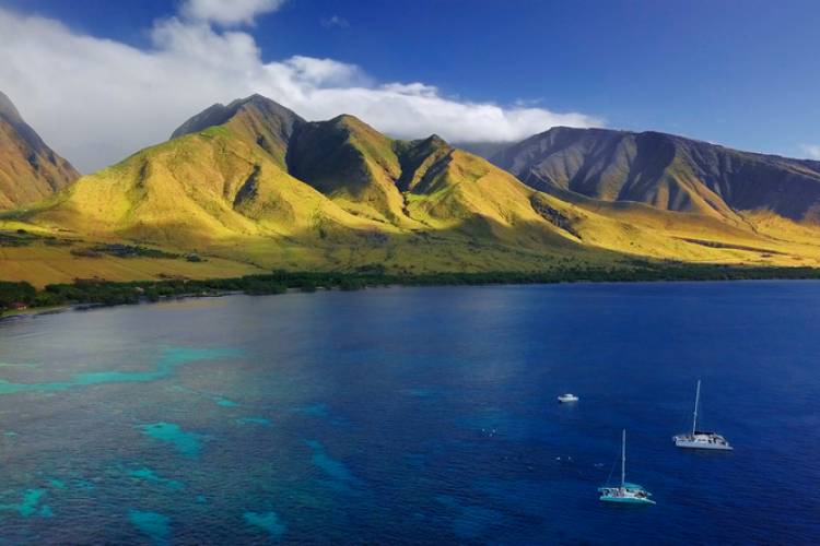 Aerial view of the west coast of Maui with visible coral reef, sailing boats, and green mountain in the background.
