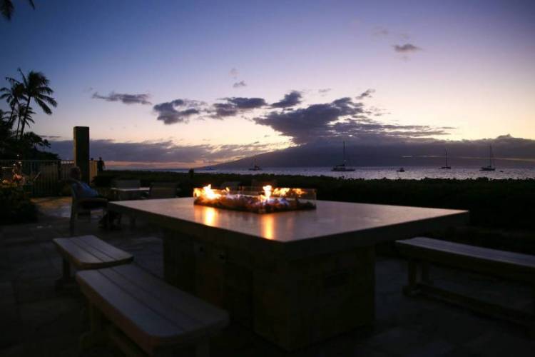 Luxury stay at The Whaler in Kaʻanapali, Maui, featuring an outdoor seating area with a table-centered fireplace glowing at sunset, overlooking the ocean with warm light reflecting across the water.