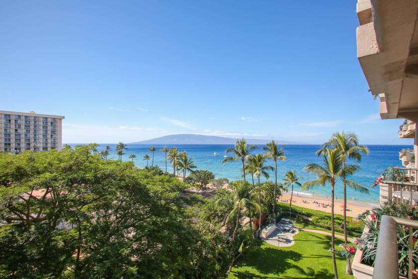Stunning view of Kaanapali’s shoreline from private lanai at The Whaler. Palm trees are in the foreground and an island rises from the sea in the distance.