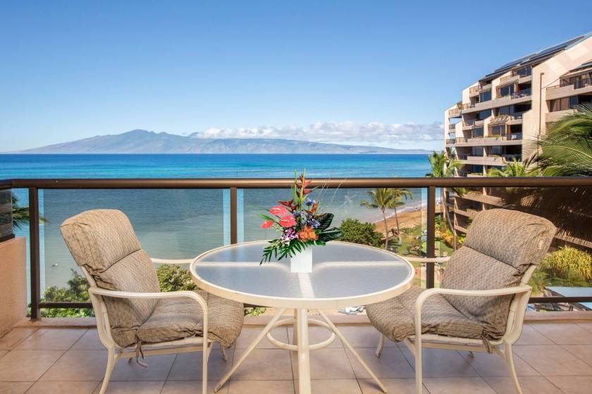 Private lanai featuring outdoor table with two chairs, overlooking view of the ocean. A flower arrangement is placed on the table. Sands of Kahana resort, Maui, HI.