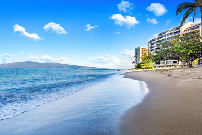 Kahana Bay stretches down the coast near Sands of Kahana resort in Maui, Hawaii. The sky is clear and the sky reflects off the wet sand.