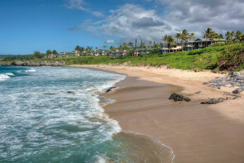 Kapalua Bay, lined with oceanfront Maui Condos and residential buildings. Palm trees sway in the background and the waves meet the shore in the foreground.