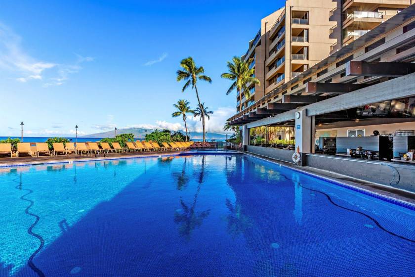 Sands of Kahana’s resort-style pool, lined with lounge chairs. The sky and palm trees are reflected in the water.